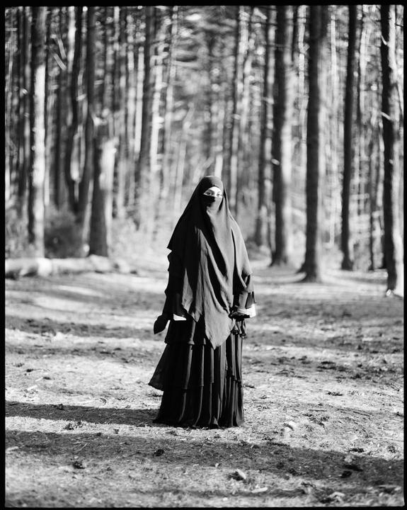 A woman wearing a niqab stands in a forest with tall trees.