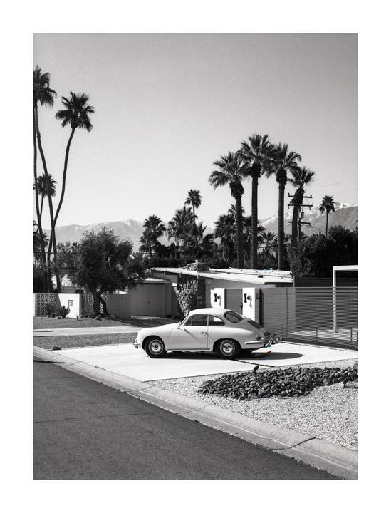 A vintage car is parked in a driveway in front of a house with palm trees and mountains in the background.