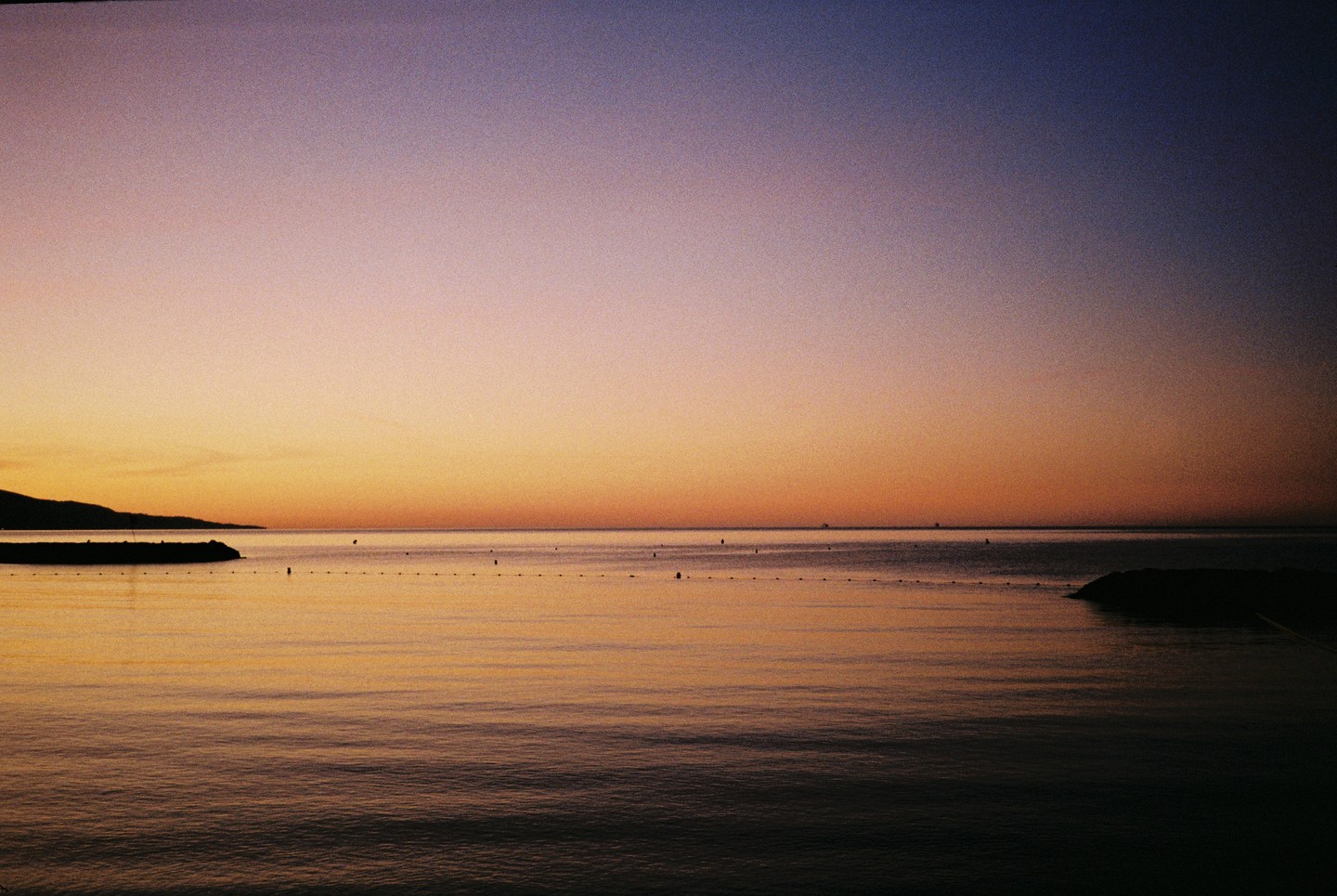 The sun sets over a calm sea with a distant coastline and a line of buoys.