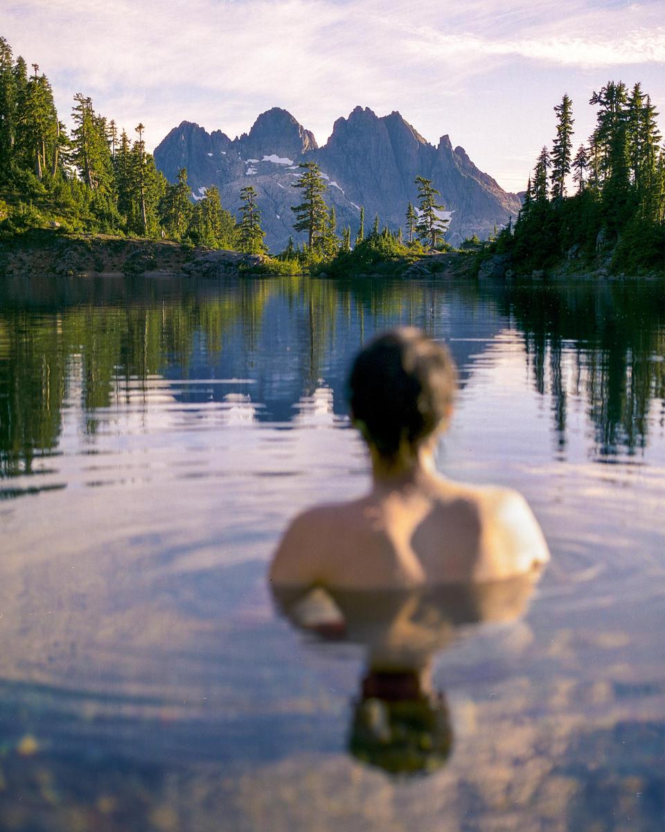 A person is in a lake looking at mountains and trees.
