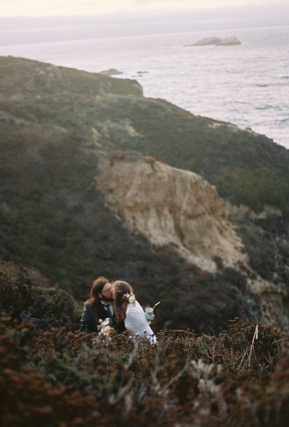 A couple kisses on a hillside overlooking the ocean.