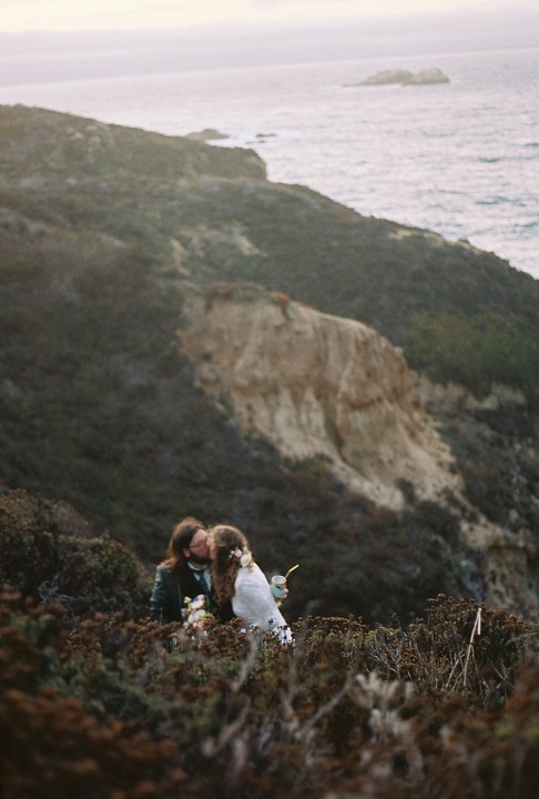 A couple kisses on a hillside overlooking the ocean.