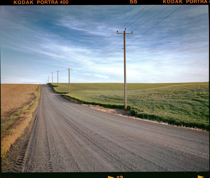 A dirt road stretches into the distance, flanked by green and brown fields under a blue sky with power lines running alongside.
