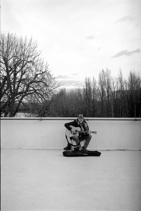 A man wearing a hat and scarf sits on a stool and plays an acoustic guitar on a rooftop.