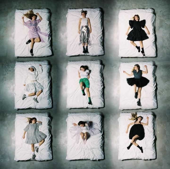 Nine women lie on nine separate beds arranged in a grid pattern on a concrete floor.