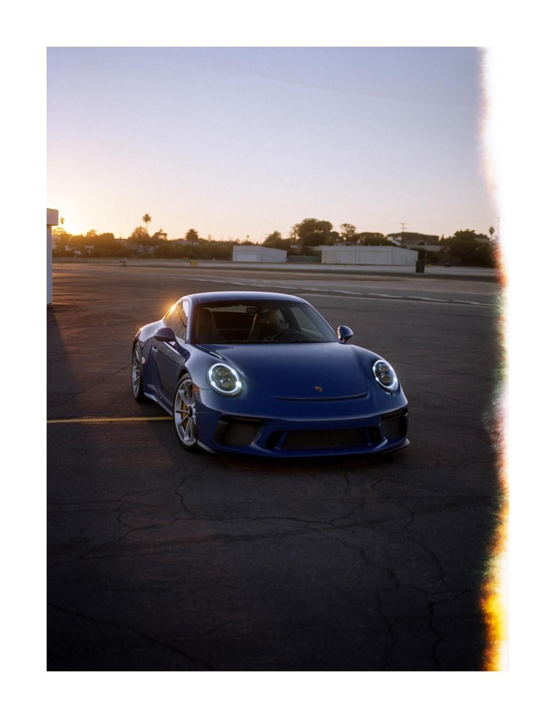 A blue sports car is parked on an asphalt lot with the setting sun in the background.
