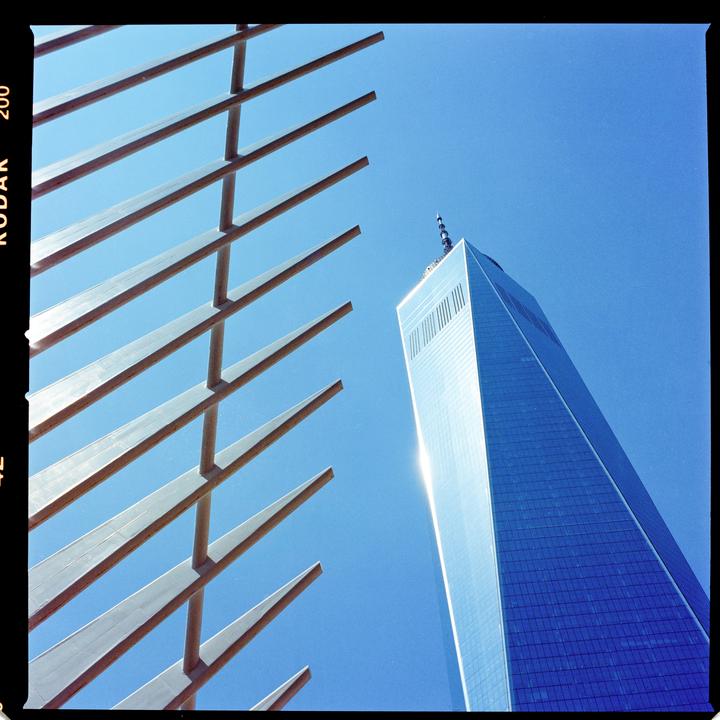 A low-angle view shows a modern skyscraper and a slatted structure against a clear blue sky.