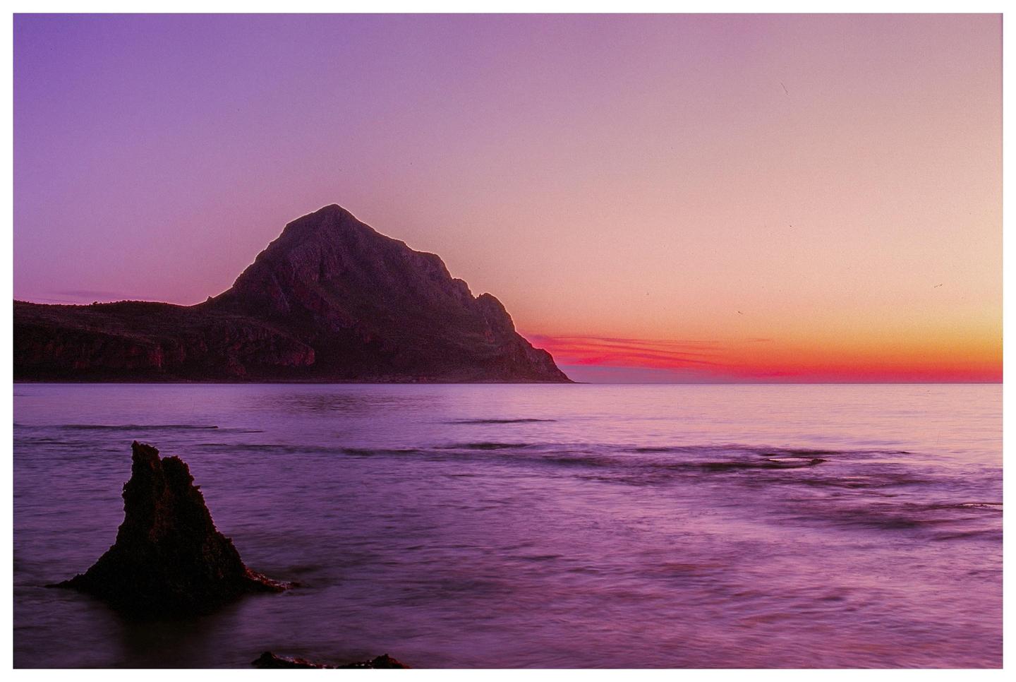 A large mountain rises from the sea under a colorful sunset sky.