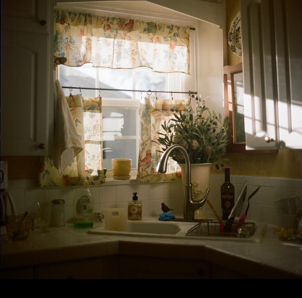 Sunlight streams through a kitchen window with patterned curtains, illuminating a sink filled with various items.