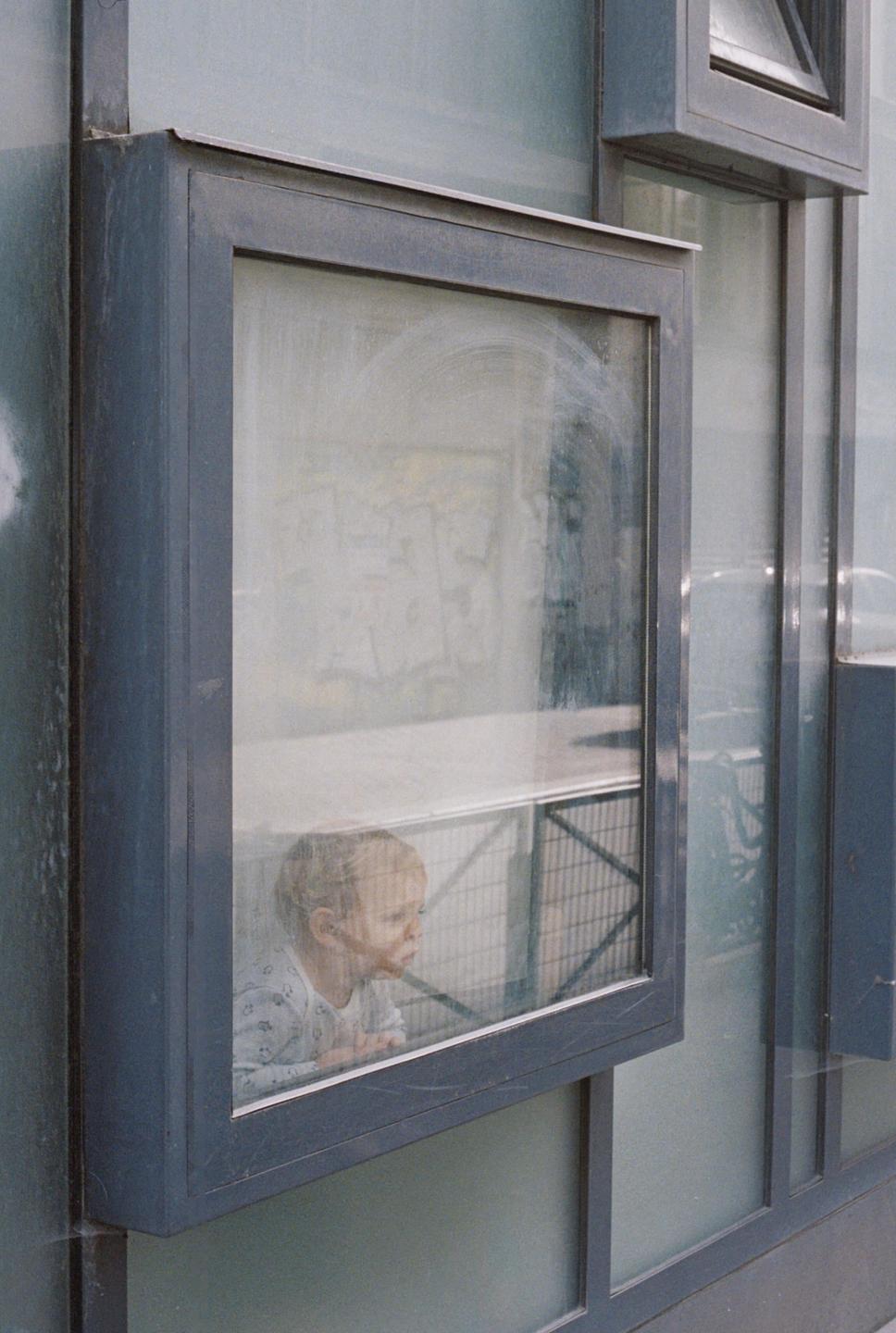 A child looks out a window in Paris, with reflections visible on the glass.