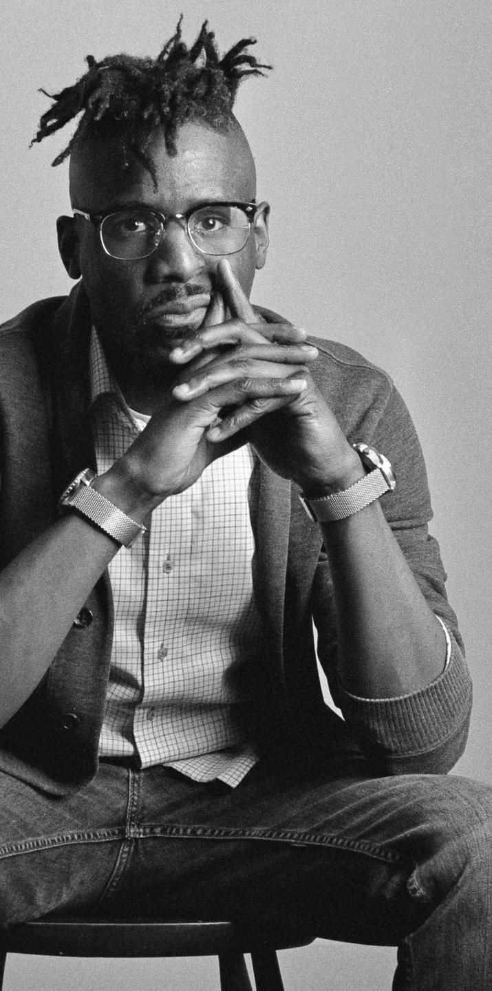 A man with dreadlocks and glasses sits with his hands clasped in front of him.