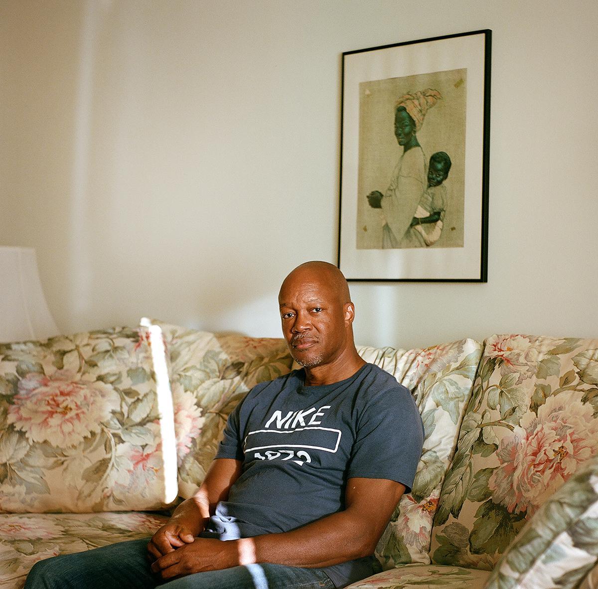A man sits on a floral couch with a framed picture of a woman and child on the wall behind him.