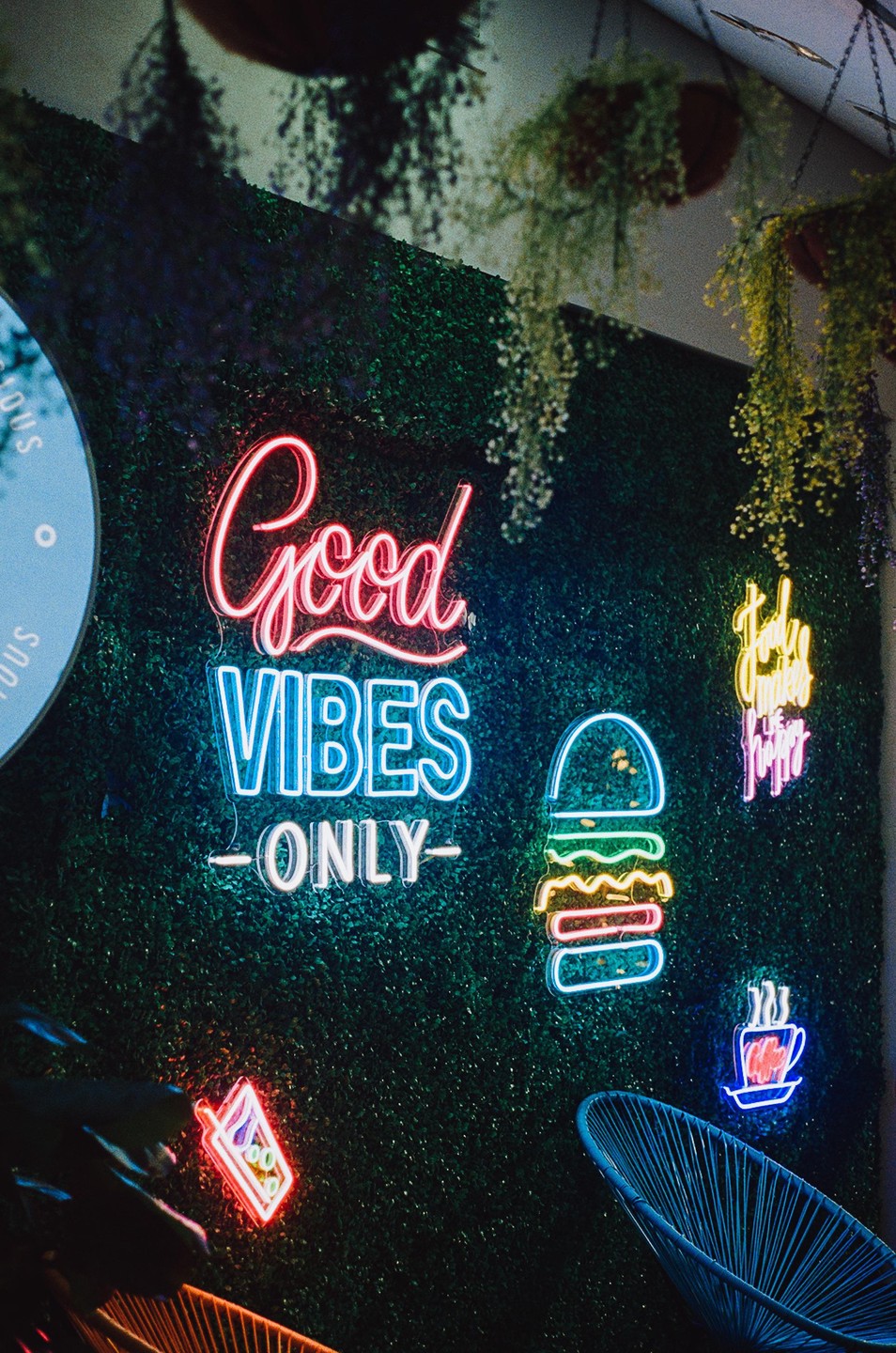 Neon signs that say "Good Vibes Only" and depict a burger, a drink, and a coffee cup are displayed on a green wall with hanging plants.