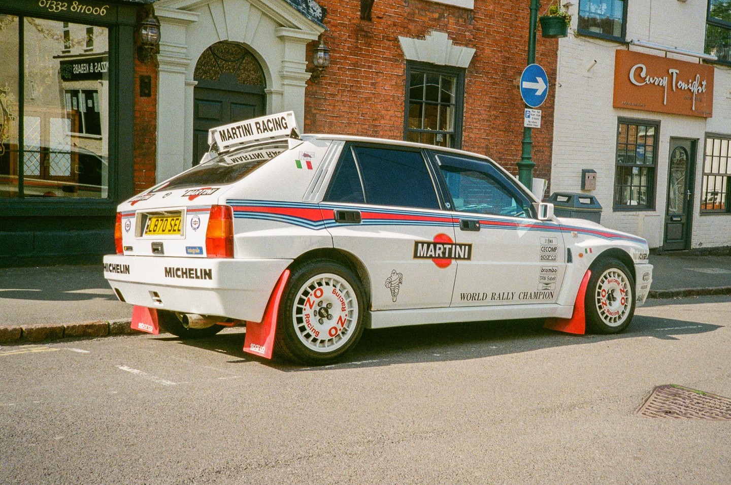 A white Martini Racing rally car is parked on a street in front of brick buildings.