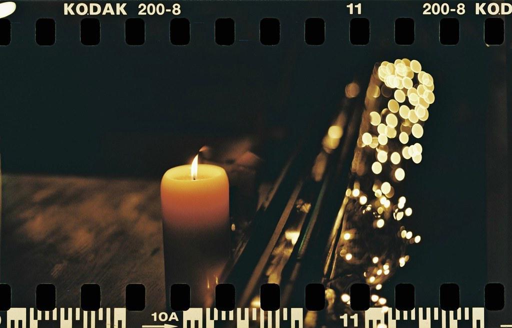 A lit candle stands next to a string of blurred lights in a dark setting.