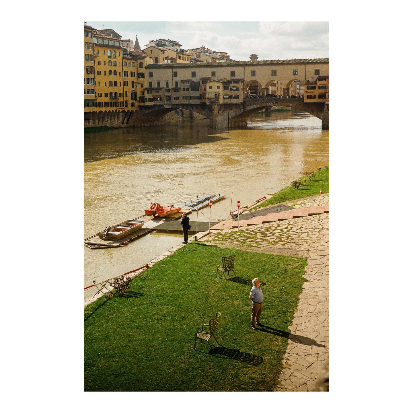 Two men stand on a grassy riverbank with a historic bridge and buildings in the background.