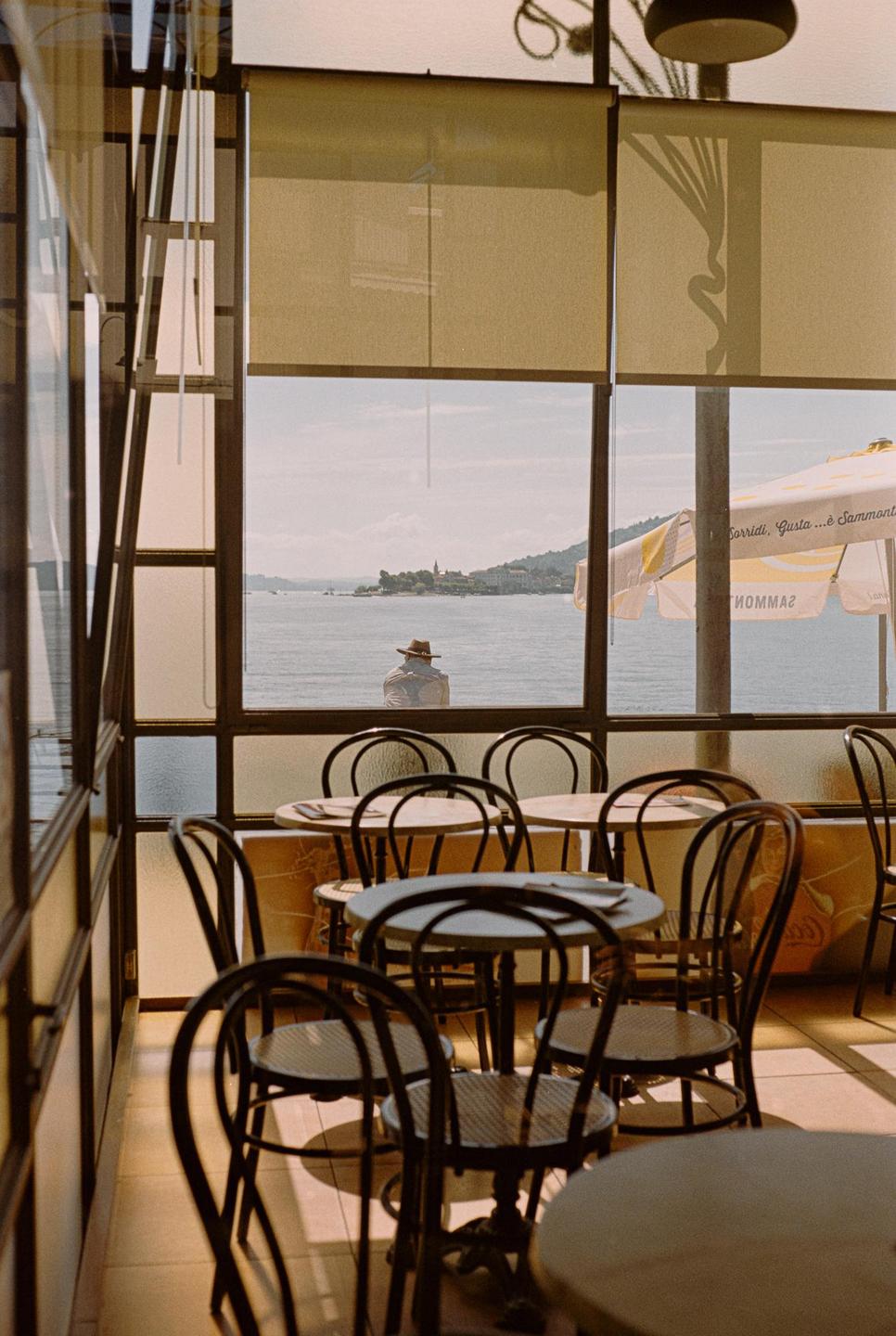A person in a hat sits by a lake, viewed from inside a cafe with empty tables and chairs.