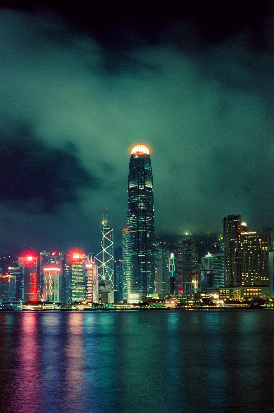 A cityscape at night shows a tall skyscraper with a glowing orange top, surrounded by other illuminated buildings, with their reflections visible in the water below.
