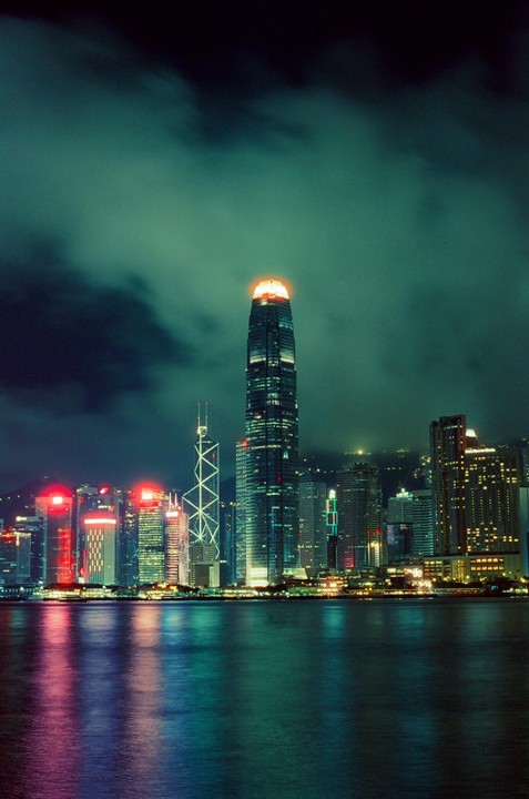 A cityscape at night shows a tall skyscraper with a glowing orange top, surrounded by other illuminated buildings, with their reflections visible in the water below.