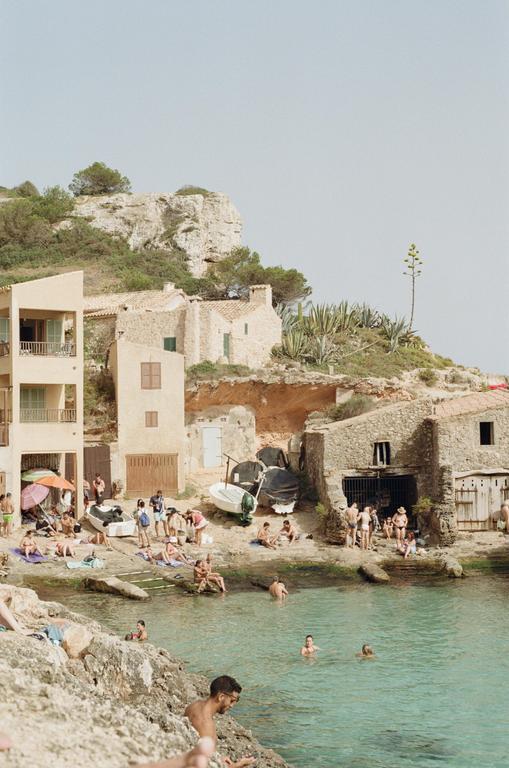 A group of people are swimming and relaxing on a rocky beach with buildings and cliffs in the background.