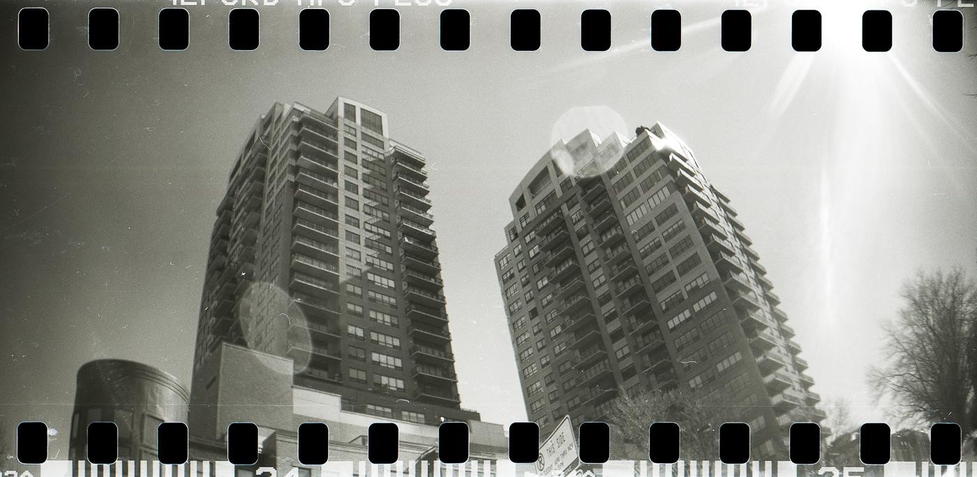 Two tall buildings with many balconies stand against a bright sky with sun flare and visible film sprockets.