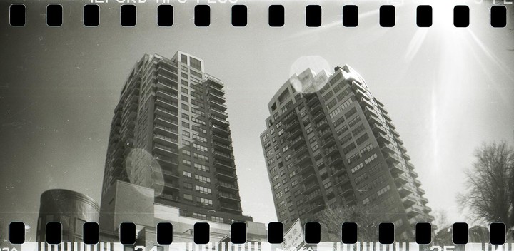 Two tall buildings with many balconies stand against a bright sky with sun flare and visible film sprockets.