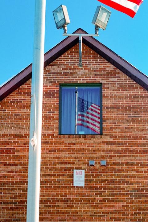 An American flag is reflected in a window on a brick building with a flagpole and lights in the foreground.