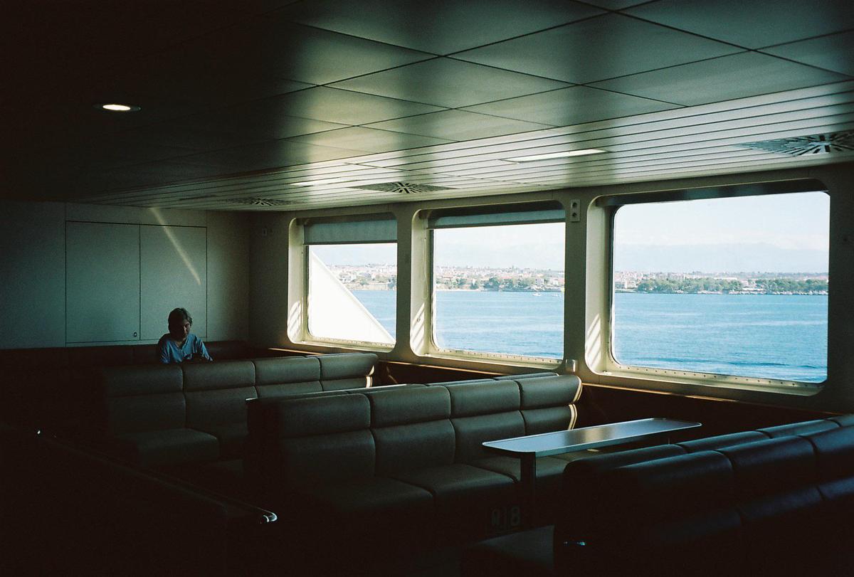 A man sits alone in a dark ferry cabin with large windows looking out onto a body of water and a distant city.