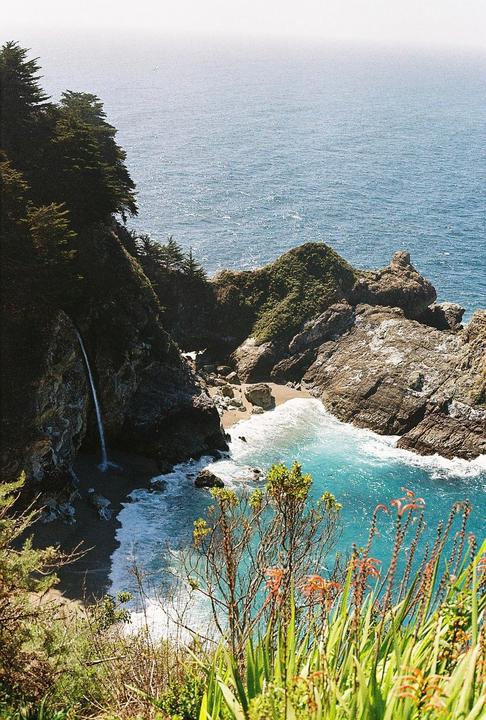 A waterfall cascades onto a small beach next to a turquoise ocean inlet surrounded by rocky cliffs and green foliage.
