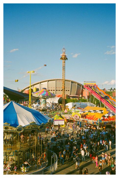 A large crowd of people gathers around various rides at a fairground under a clear blue sky.