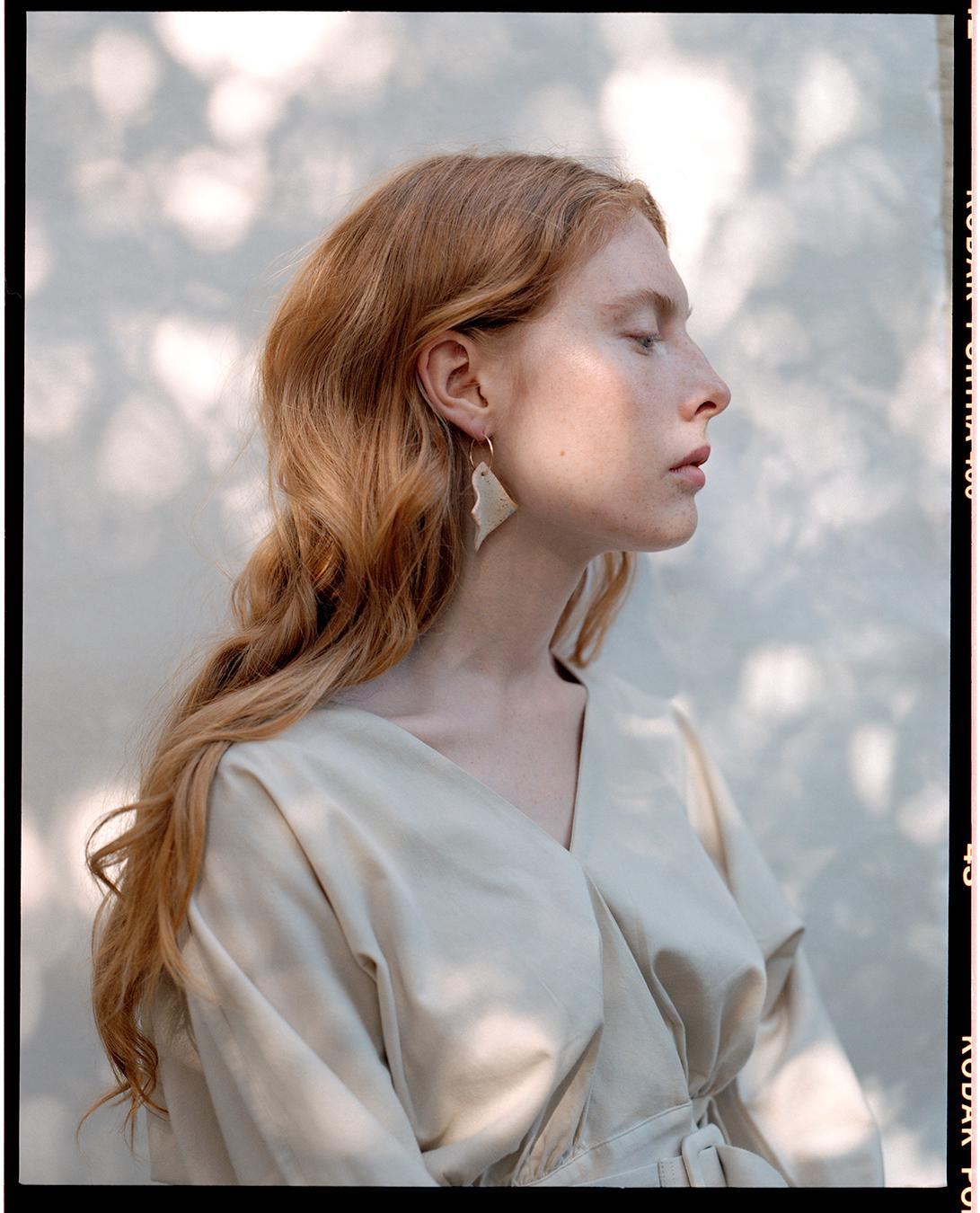 A woman with long red hair and an earring is shown in profile against a wall with dappled light.