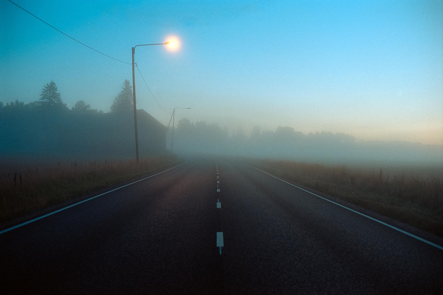 A road stretches into the distance, illuminated by a single street light in a hazy, blue-tinged landscape.