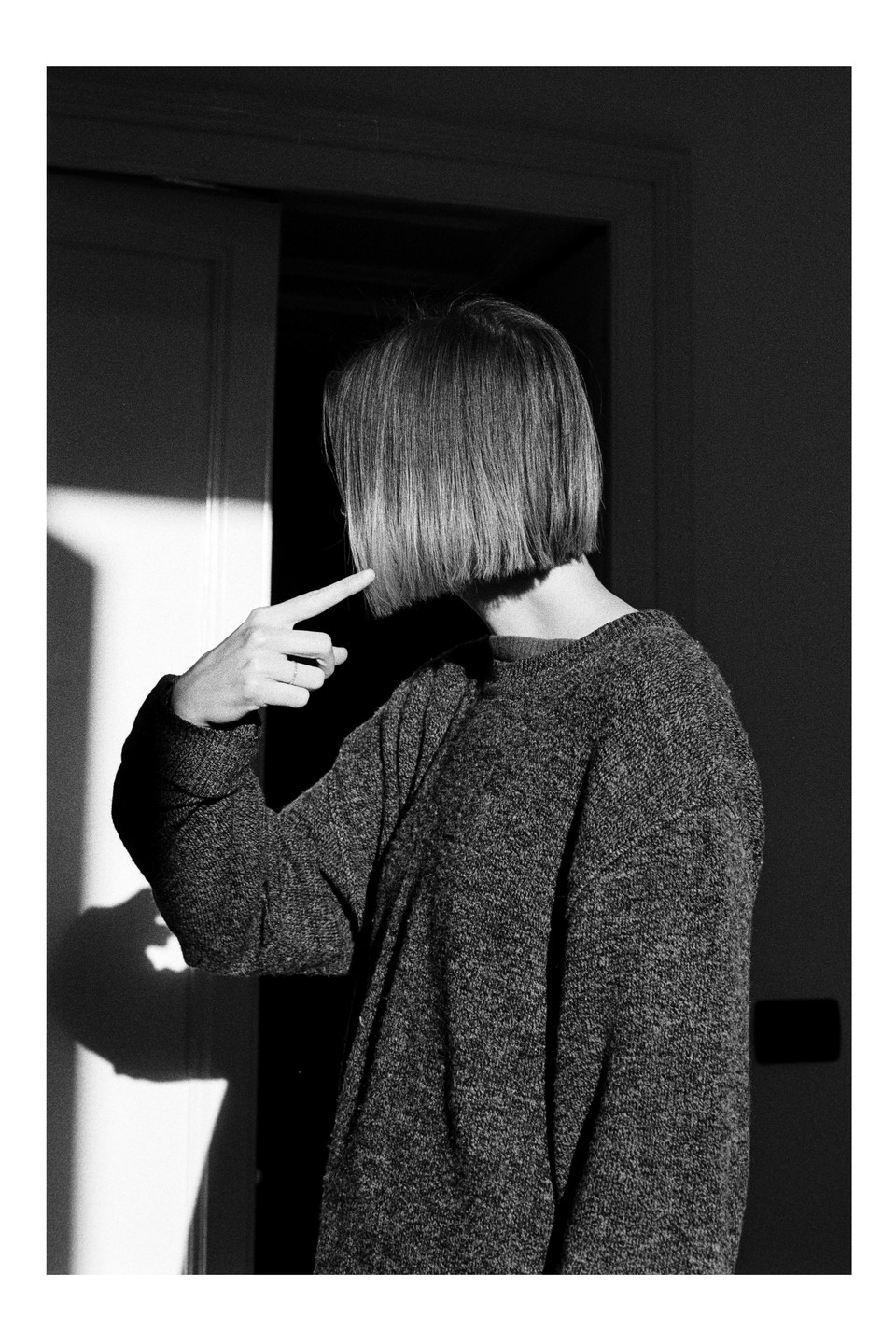 A woman with a bob haircut points to her hair, standing in a room with strong shadows.