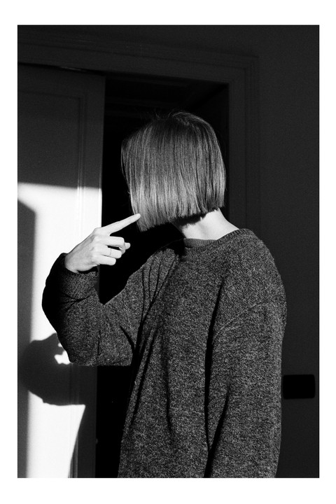 A woman with a bob haircut points to her hair, standing in a room with strong shadows.