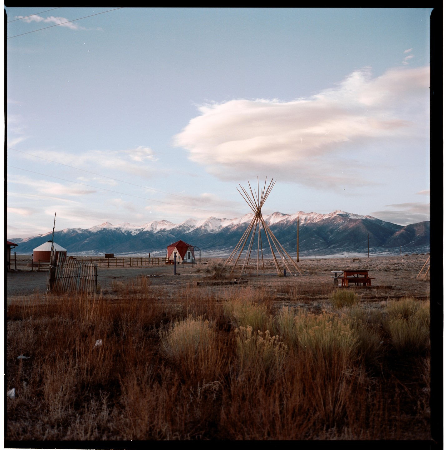 A wide-angle landscape shows a field with a teepee frame, a small red building, and a silo, with snow-capped mountains in the background under a cloudy sky.