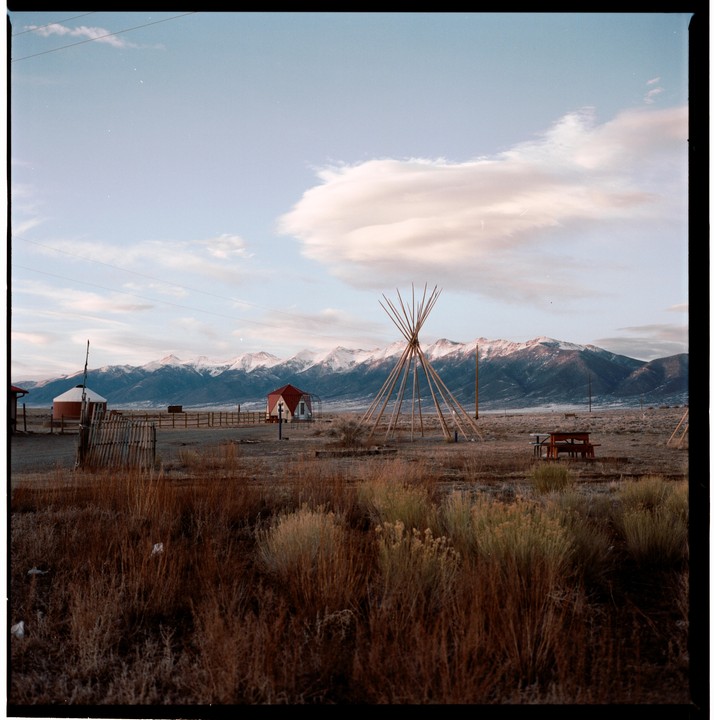 A wide-angle landscape shows a field with a teepee frame, a small red building, and a silo, with snow-capped mountains in the background under a cloudy sky.