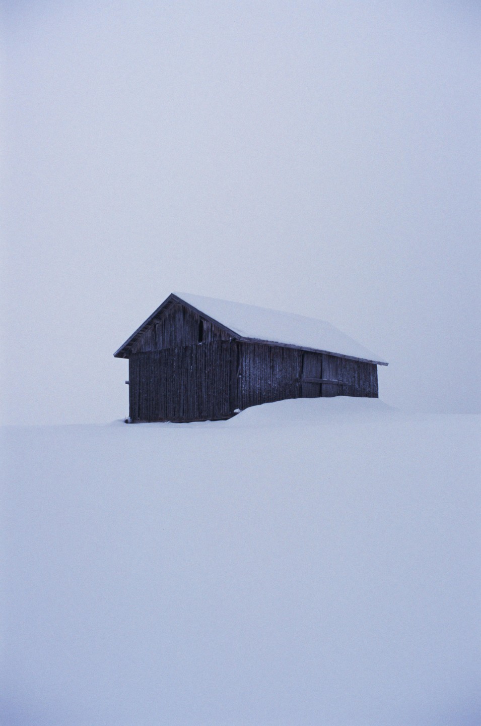A wooden barn is partially covered in snow in a vast, snowy landscape under a pale sky.