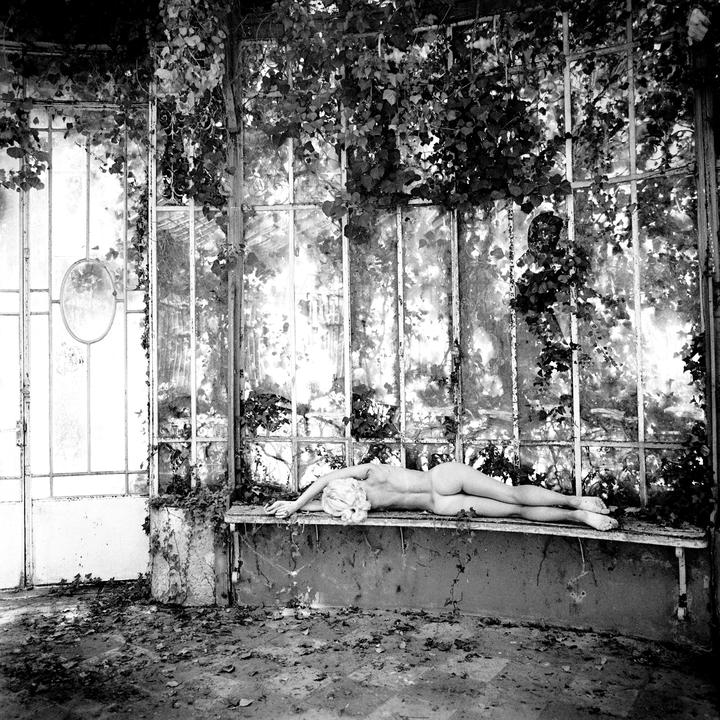 A nude woman with blonde hair lies on a bench in a greenhouse covered in vines.