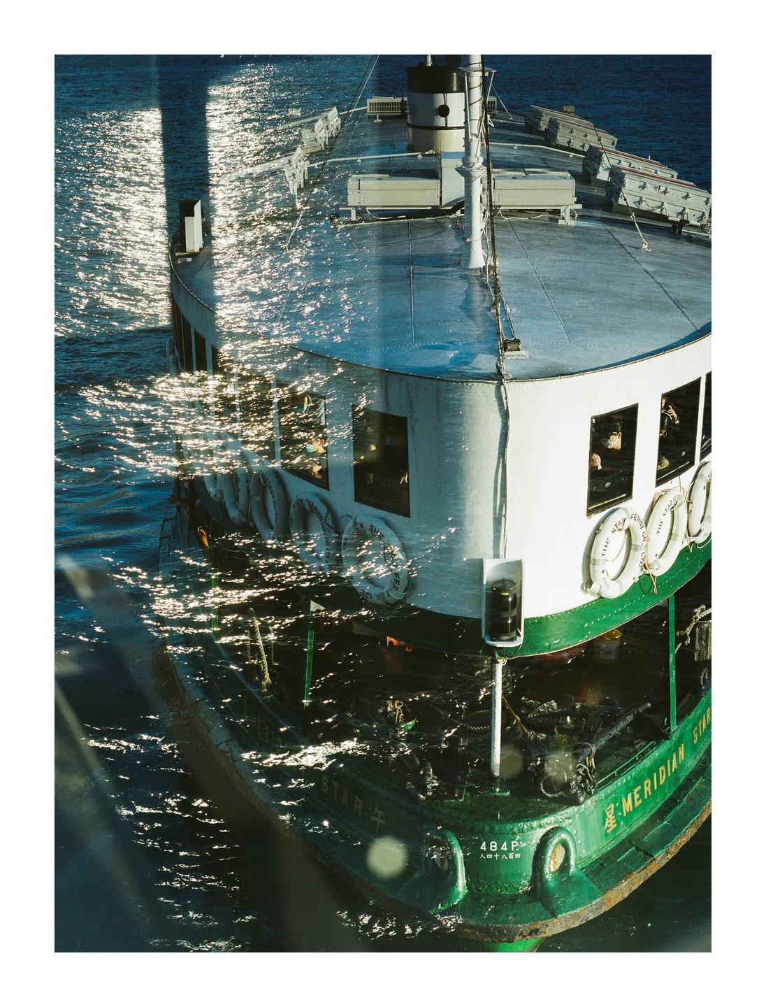 A ferry boat with a green hull and white upper deck moves through sparkling water, seen from an elevated perspective.