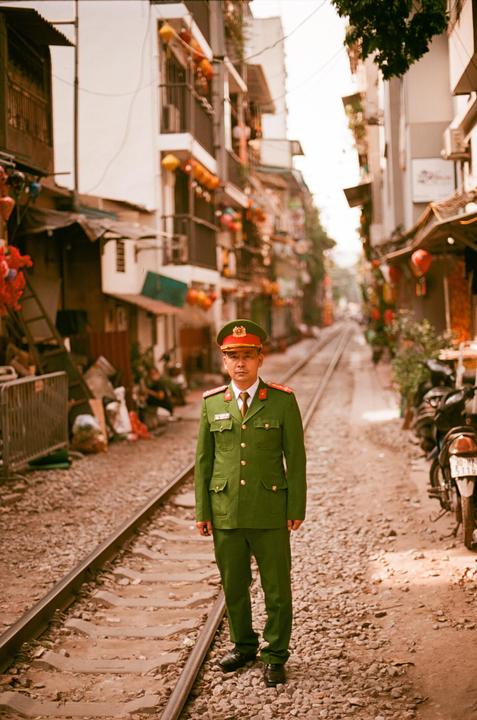 A man in a green uniform stands on train tracks in a narrow street lined with buildings.