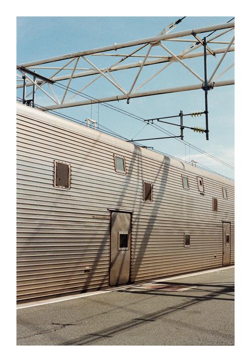 A train car with multiple small windows and doors is parked next to a platform, with overhead power lines and structures visible against a clear sky.