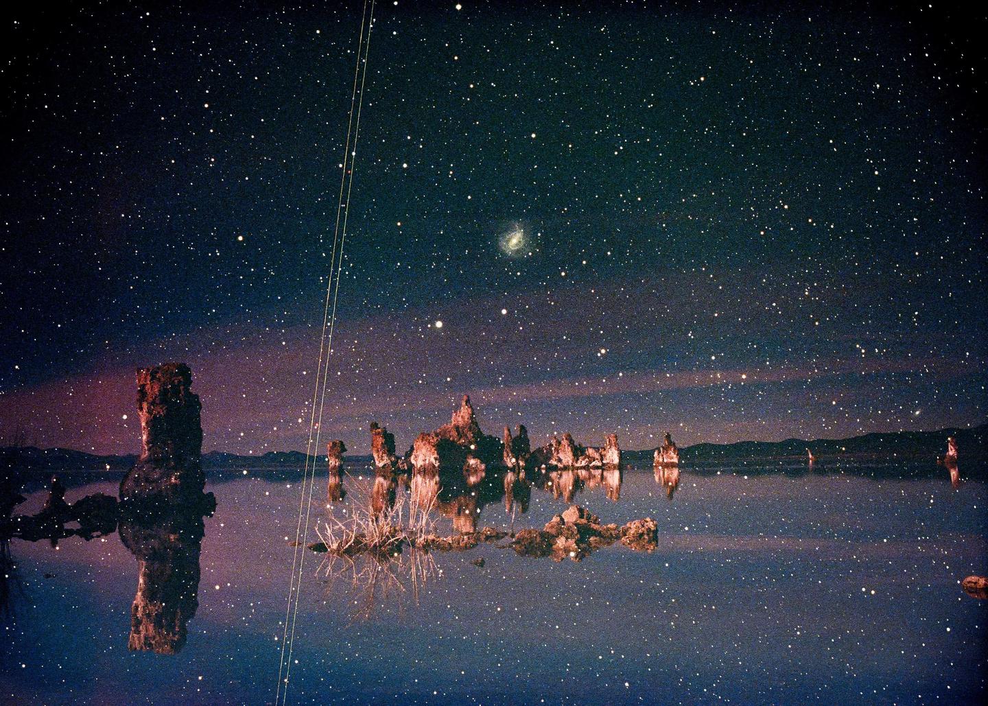 A starry night sky with a visible galaxy is reflected in the still water of Mono Lake, surrounded by tufa formations.
