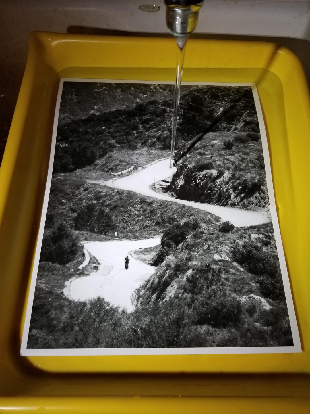 A black and white print of a winding road with a person on it is being rinsed under running water in a yellow tray.