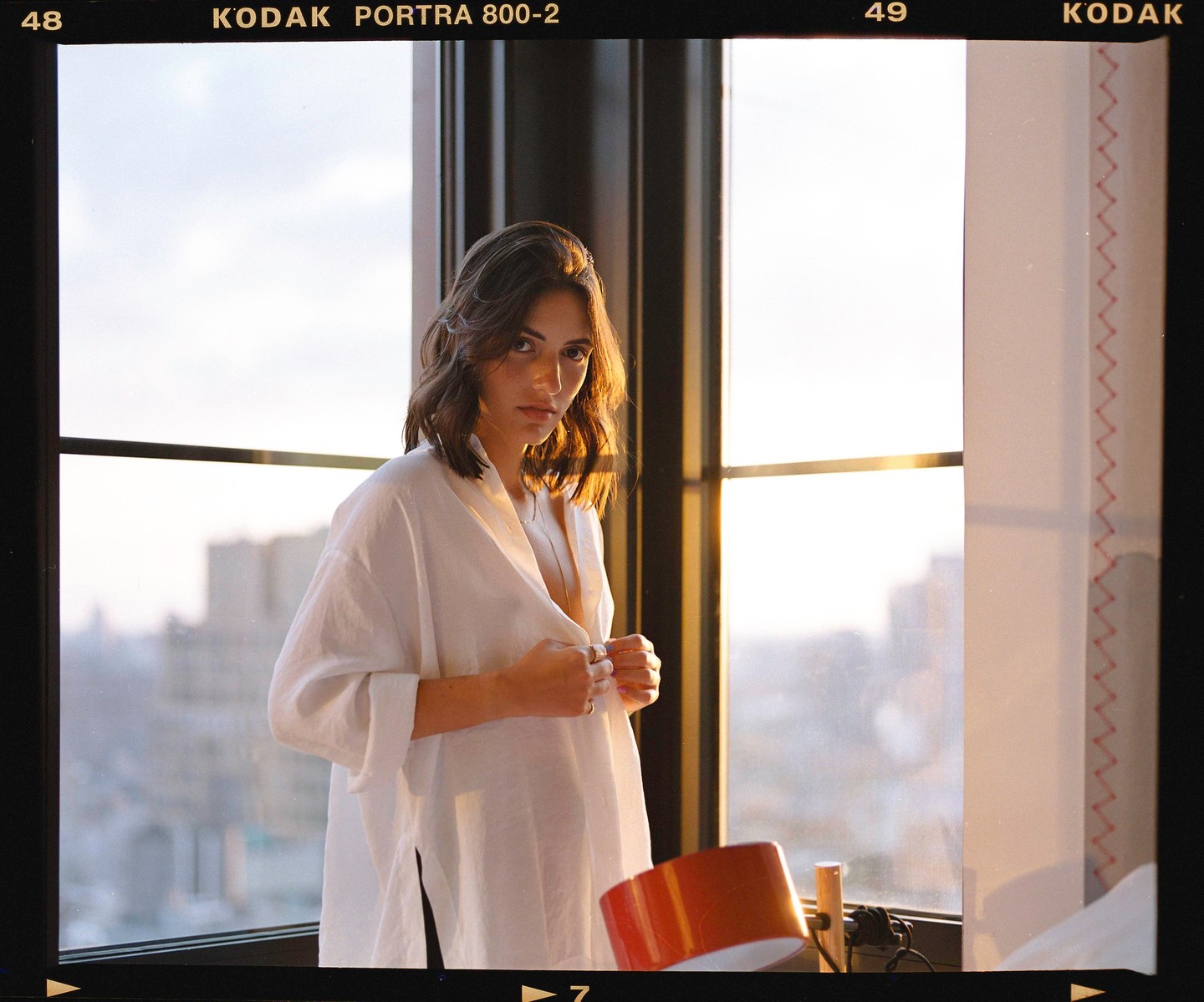 A woman in a white shirt stands by a window with a city view at sunset.
