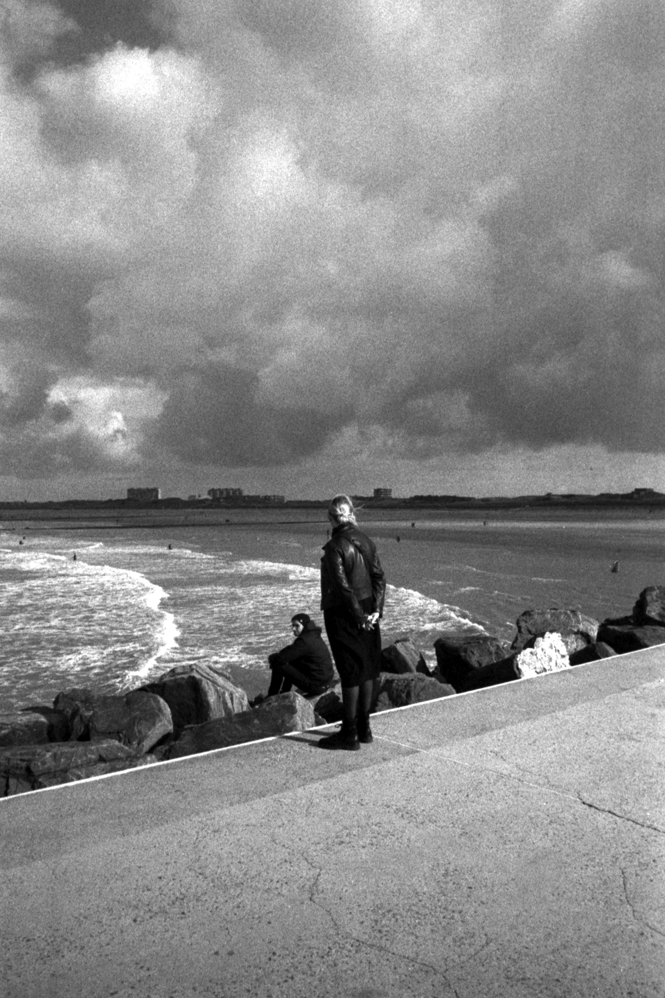 Two people stand on a rocky shore looking out at a wavy sea under a cloudy sky.