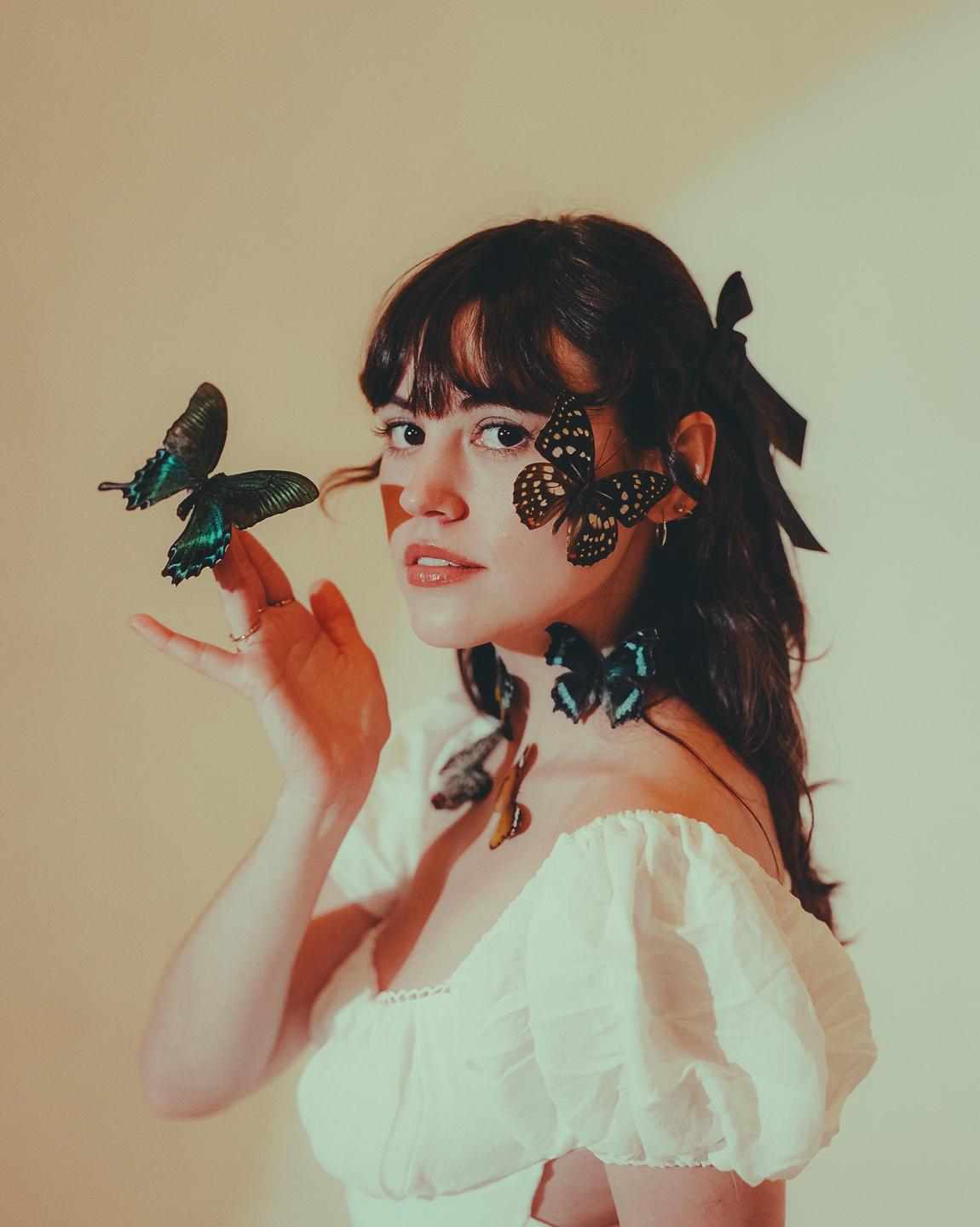 A woman with dark hair and a white top has several butterflies on her face, neck, and hand.