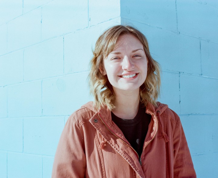 A woman with a nose ring smiles at the camera while standing in front of a blue wall.
