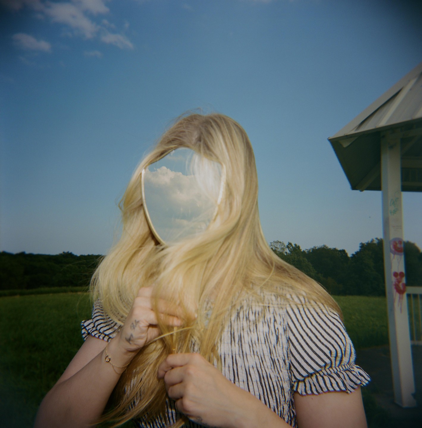 A woman with long blonde hair holds a mirror reflecting the sky in front of her face.