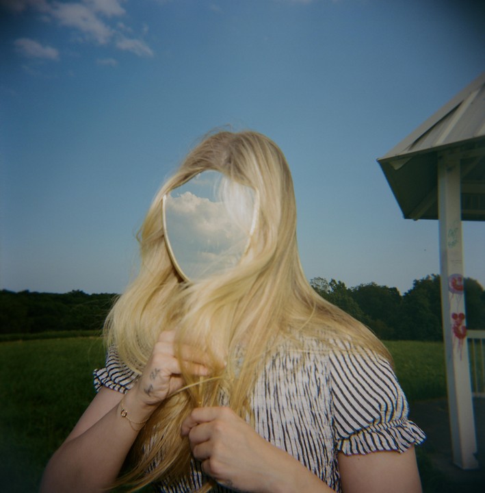 A woman with long blonde hair holds a mirror reflecting the sky in front of her face.