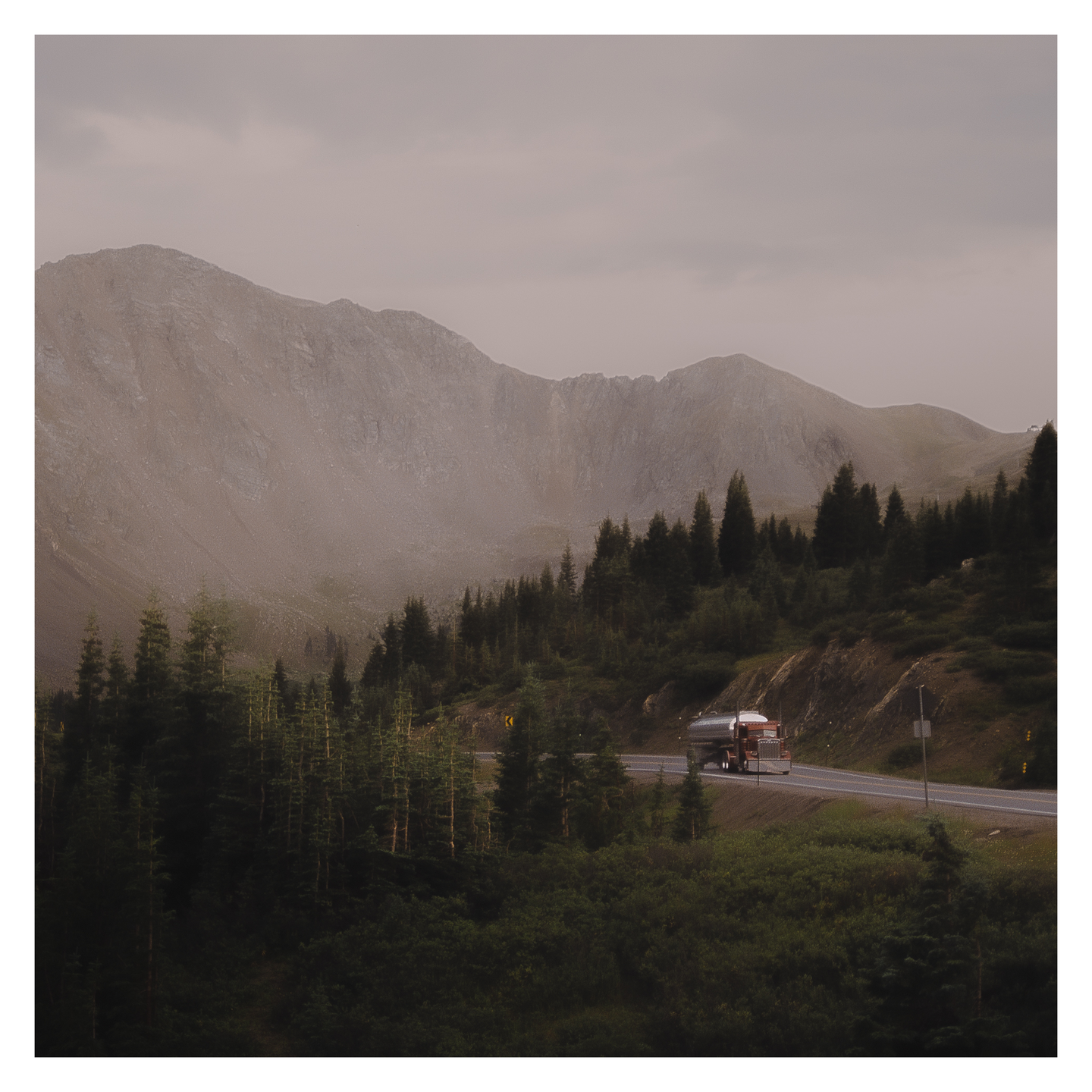 A truck drives along a winding road through a mountainous landscape with pine trees under a cloudy sky.